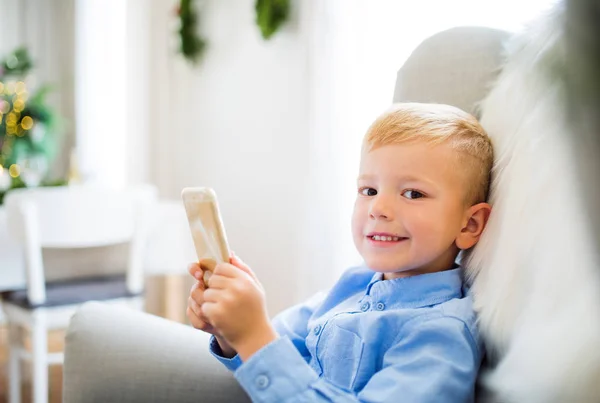 Маленький хлопчик зі смартфоном сидить на кріслі вдома в різдвяний час . — стокове фото