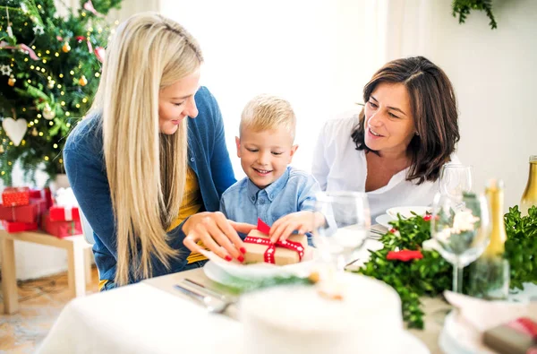 Um menino pequeno com presente e mãe e avó em casa na época do Natal . — Fotografia de Stock