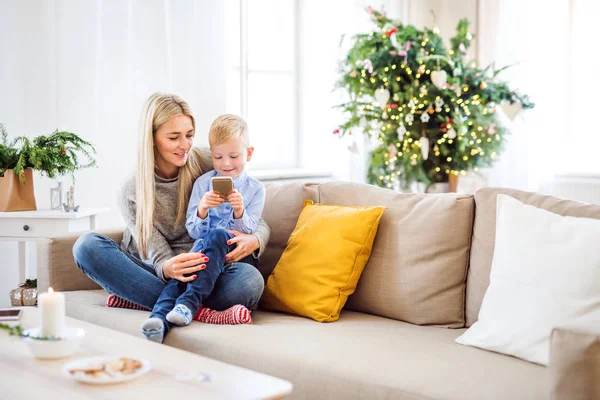 Uma mãe e um menino pequeno com smartphone sentado em um sofá em casa na época do Natal . — Fotografia de Stock