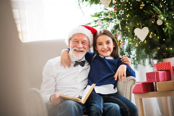 Uma menina pequena e seu avô com chapéu de Papai Noel lendo um livro na época do Natal . — Fotografia de Stock