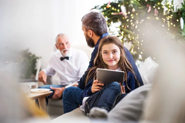 Niña pequeña con la tableta y su padre y abuelo sentado en un sofá en tiempo de Navidad . —  Fotos de Stock