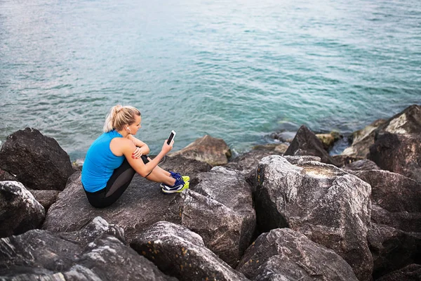 Joven corredora deportiva con auriculares y smartphone sentado en las rocas por mar . —  Fotos de Stock