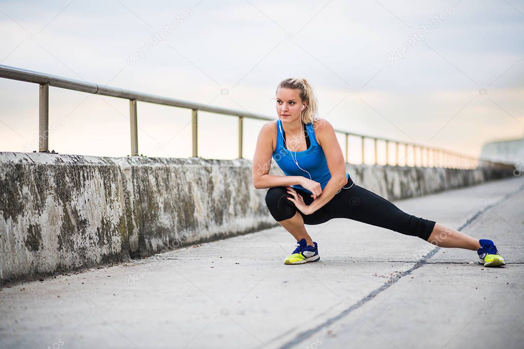 Young sporty woman runner with earphones stretching by the sea outside.