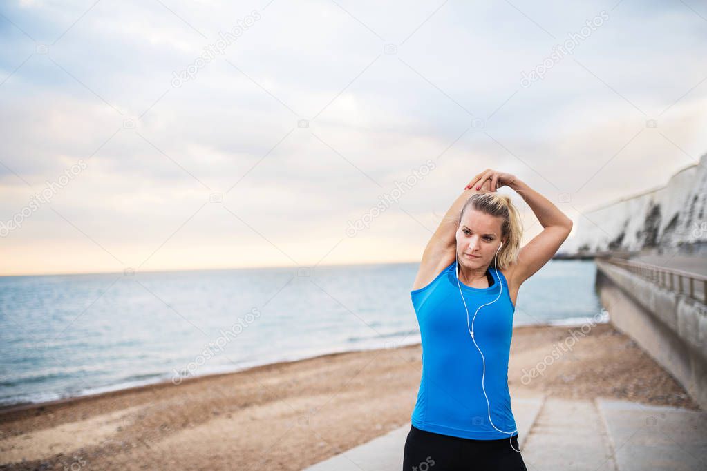 Young sporty woman runner with earphones standing on the beach outside, stretching.