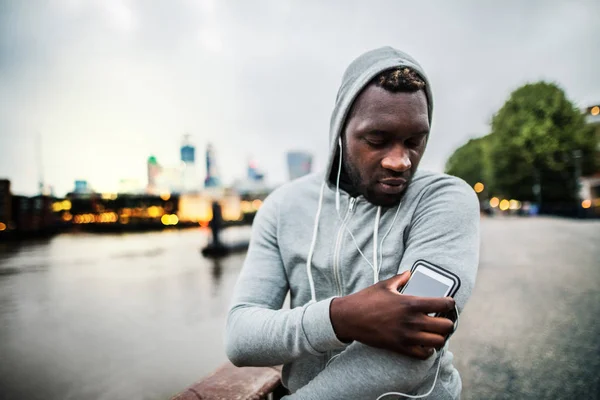 Fekete férfi futó okostelefon-karszalag a hídon, egy város, pihenő. — Stock Fotó