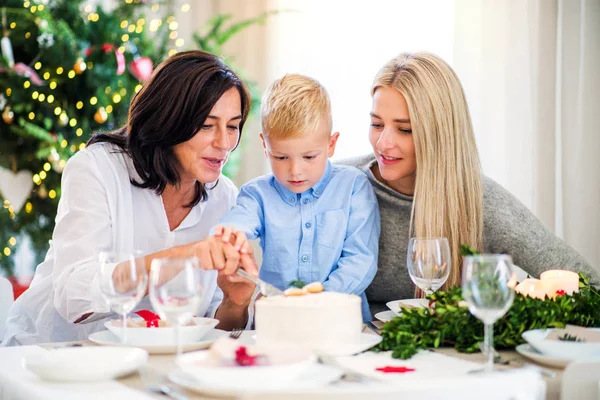 Um menino pequeno com mãe e avó cortando um bolo na época do Natal . — Fotografia de Stock