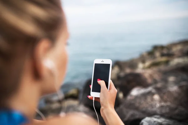 Sportig kvinna löpare med hörlurar och smartphone sitter på klipporna vid havet. — Stockfoto