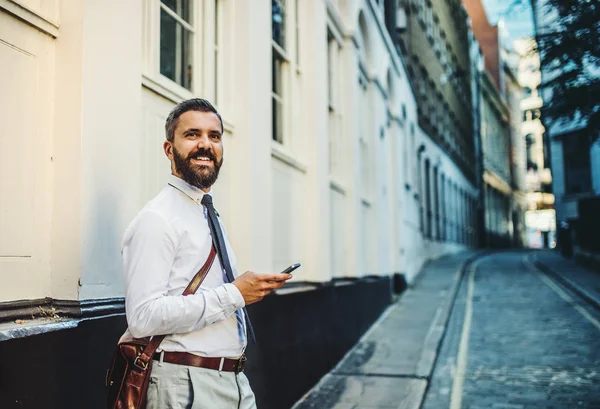 Hipster hombre de negocios de pie en la calle en Londres, sosteniendo el teléfono inteligente . — Foto de Stock