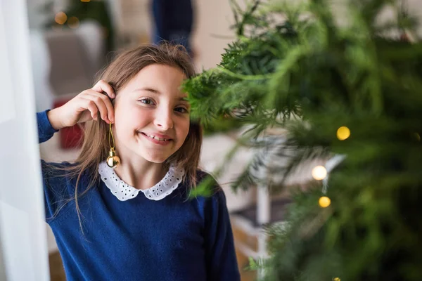 Small girl standing by a Christmas tree at home, a ball ornament on her ear. Copy space. — Stock Photo, Image