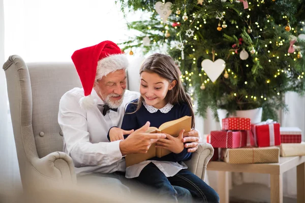 A small girl and her grandfather with Santa hat and a book at Christmas time. — Stock Photo, Image