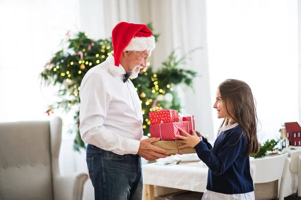 Um homem sênior com um chapéu de Papai Noel dando presentes para uma menina pequena na época do Natal . — Fotografia de Stock