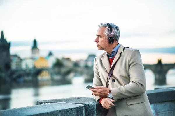 Hombre de negocios maduro con auriculares y teléfono inteligente de pie junto al río en la ciudad . — Foto de Stock