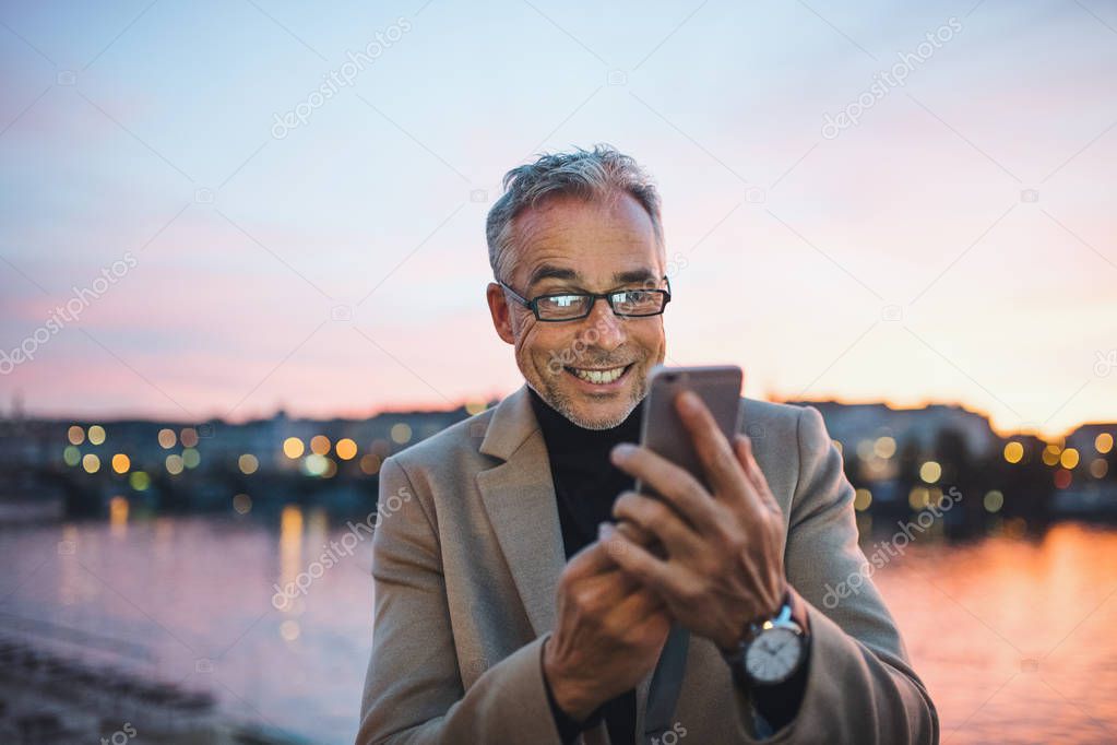Mature businessman with smartphone standing by river in Prague city, taking selfie.