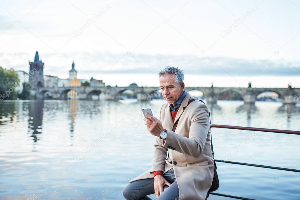 Mature businessman with smartphone sitting by river Vltava in Prague city.