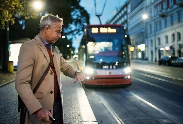 Érett üzletember vár egy villamos este bőrönddel. — Stock Fotó