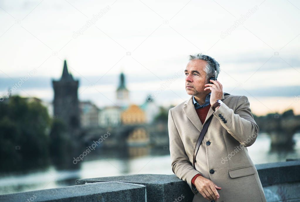 Mature businessman with headphones standing by river in city, listening to music.