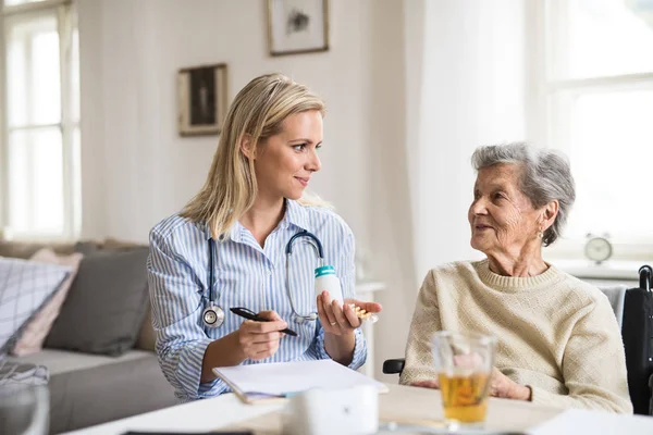 Un visitatore di salute che spiega a una donna anziana sulla sedia a rotelle come prendere pillole . — Foto Stock