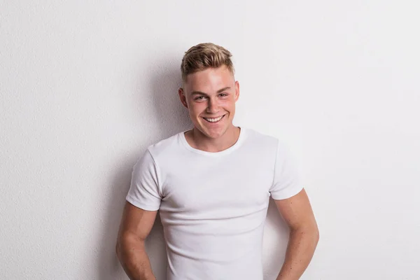 Portrait of a young man in white T-shirt in a studio, leaning against wall. — Stock Photo, Image