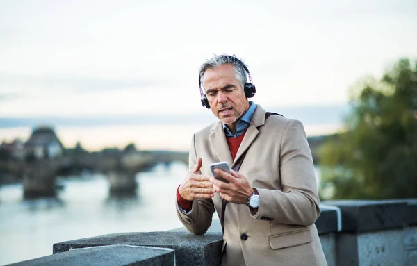 Uomo d'affari maturo con cuffie e smartphone in piedi lungo il fiume, cantando . — Foto Stock