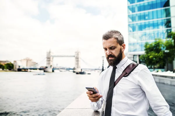 Hipster businessman with smartphone standing by the river in London, texting. — Stock Photo, Image