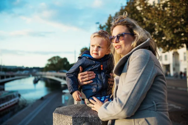 Jonge moeder met een zoon van de peuter permanent buitenshuis door de rivier in stad. — Stockfoto