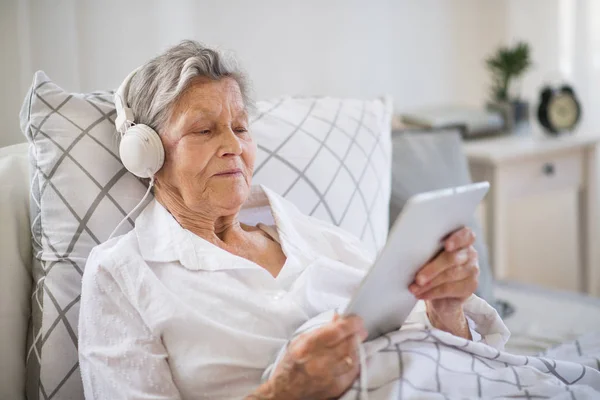 Kranke Seniorin mit Kopfhörer und Tablet zu Hause oder im Krankenhaus im Bett liegend. — Stockfoto