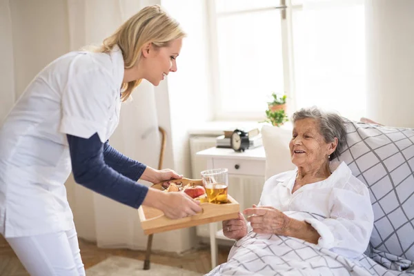 Um visitante de saúde trazendo café da manhã para uma mulher idosa doente deitada na cama em casa . — Fotografia de Stock