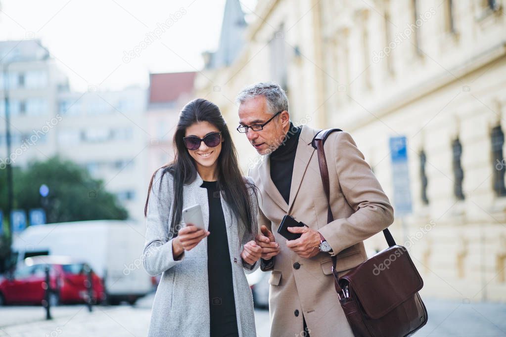 Man and woman business partners with smartphone standing outdoors in city, talking.