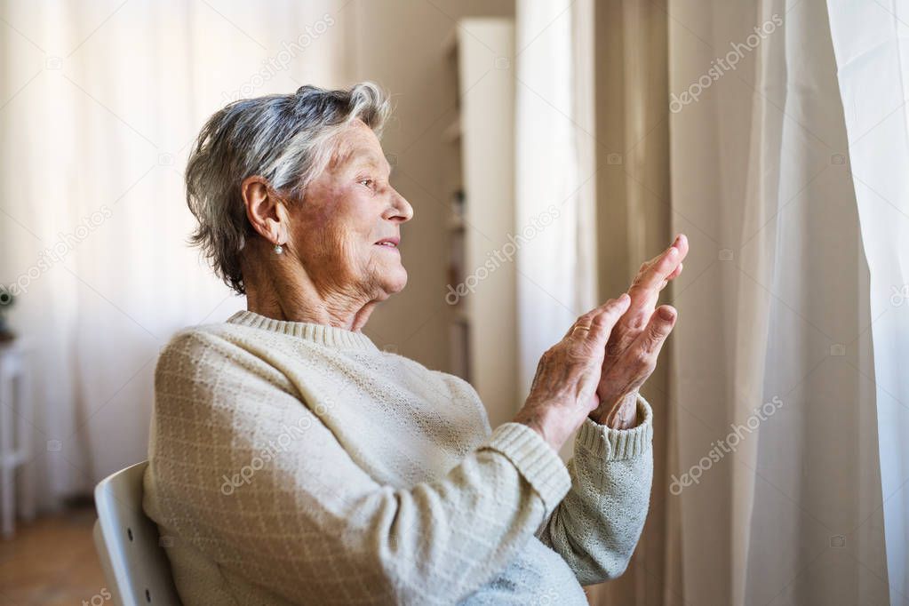 A portrait of a senior woman sitting at home, looking out of a window.