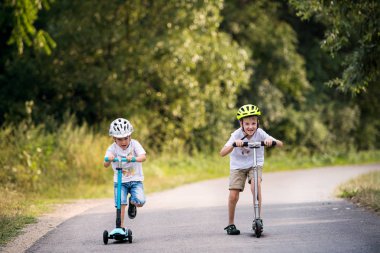 Bir yaz günü parkta bir yolda scooter sürme bir kask ile iki küçük çocuklar.