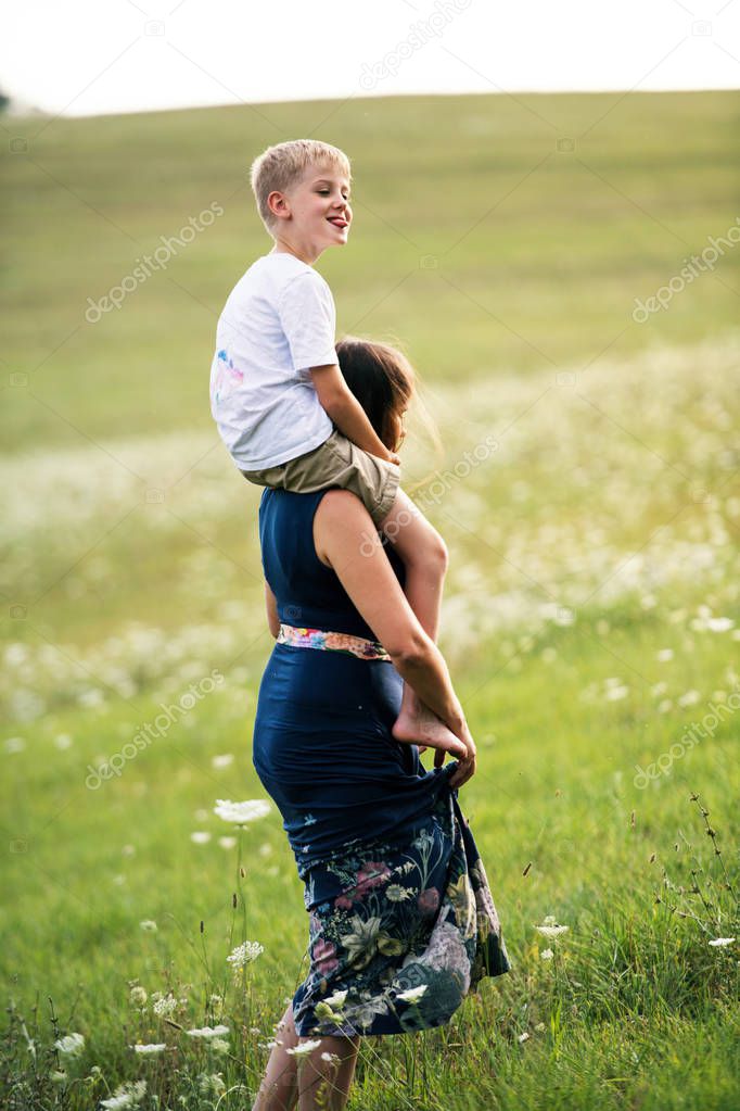 A young mother giving piggyback ride to small son in nature on a summer day.