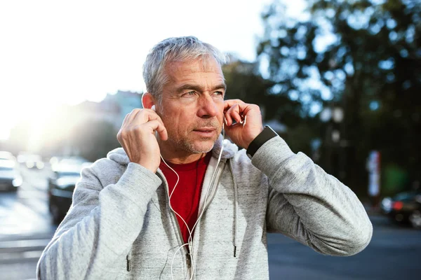 Ett porträtt av en aktiv mogen man med hörlurar som står utomhus i staden. — Stockfoto