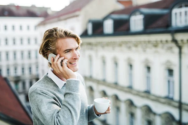 Νέος άνθρωπος με smartphone στέκεται σε ένα μπαλκόνι στην πόλη, κάνοντας ένα τηλεφώνημα. — Φωτογραφία Αρχείου