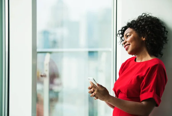 Un retrato de la mujer de pie junto a la ventana, sosteniendo el teléfono inteligente . — Foto de Stock