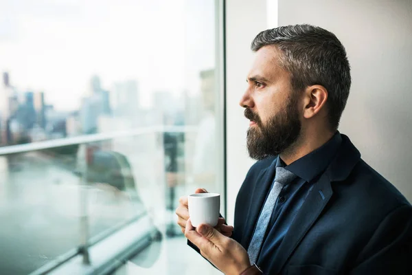 Un retrato de hombre de negocios con una taza de café mirando por una ventana en una oficina . — Foto de Stock