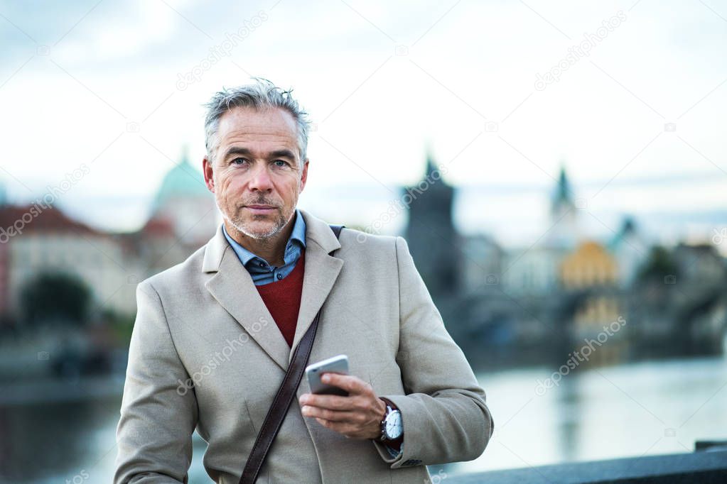 Mature businessman with smartphone standing by river Vltava in Prague city.