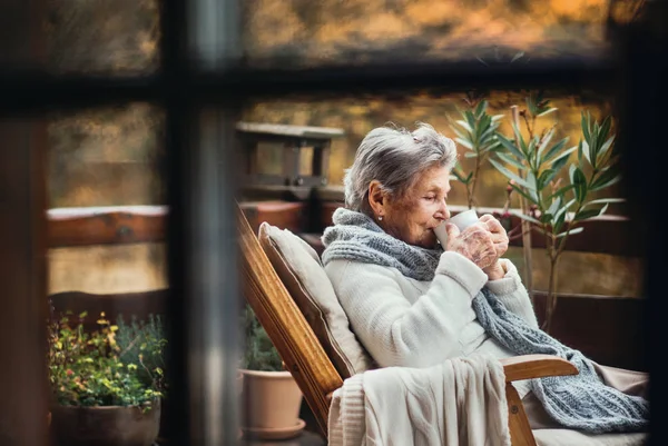 Eine ältere Frau sitzt an einem Herbsttag draußen auf einer Terrasse und trinkt Kaffee. — Stockfoto