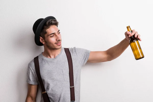 Un joven confiado con sombrero en un estudio, sosteniendo una botella de cerveza vacía . — Foto de Stock