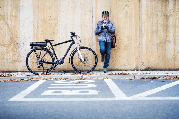 Aktiver Senior mit Elektrofahrrad steht draußen in der Stadt und nutzt Smartphone. — Stockfoto