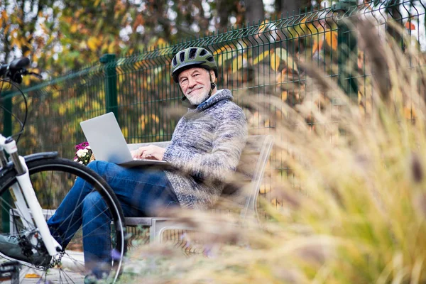 Un hombre mayor con electrobicicleta sentado en un banco al aire libre en la ciudad, usando un ordenador portátil . — Foto de Stock