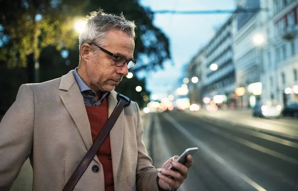 Dojrzałe biznesmen z smartphone stojący przy ulicy w godzinach wieczornych. — Zdjęcie stockowe