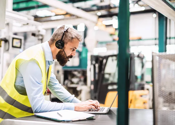 Um engenheiro industrial com fone de ouvido e laptop em uma fábrica, trabalhando . — Fotografia de Stock