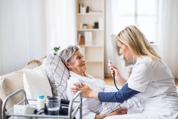 Um visitante de saúde examinando uma mulher idosa doente deitada na cama em casa com estetoscópio . — Fotografia de Stock