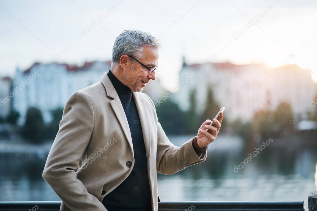 Mature businessman with smartphone standing by river in Prague city, taking selfie.