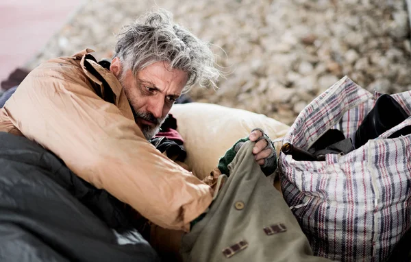 Homme mendiant sans abri couché dehors en ville, sortant quelque chose d'un sac . — Photo