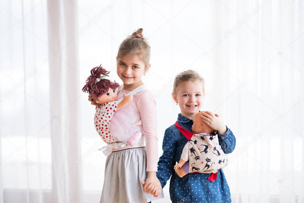 A portrait of two small girls standing and carrying dolls in baby carriers indoors.
