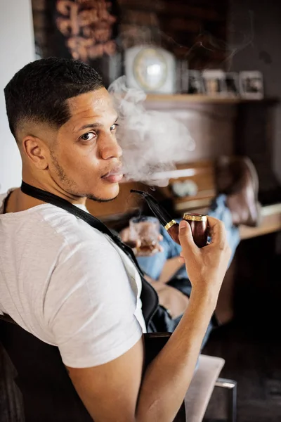Jovem hispânico haidresser e cabeleireiro sentado na barbearia, fumando um cachimbo . — Fotografia de Stock