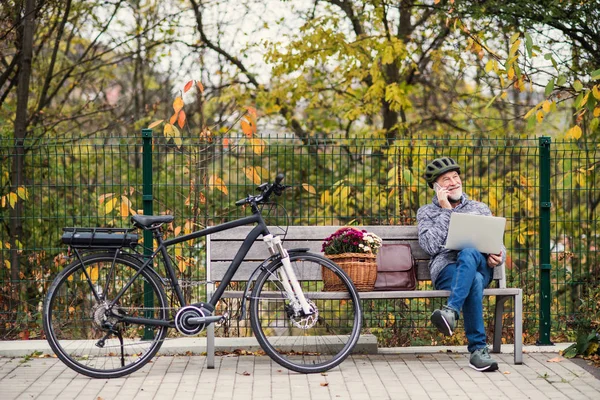 Hombre mayor con electrobicicleta sentado en un banco al aire libre en la ciudad, utilizando un ordenador portátil y un teléfono inteligente . — Foto de Stock