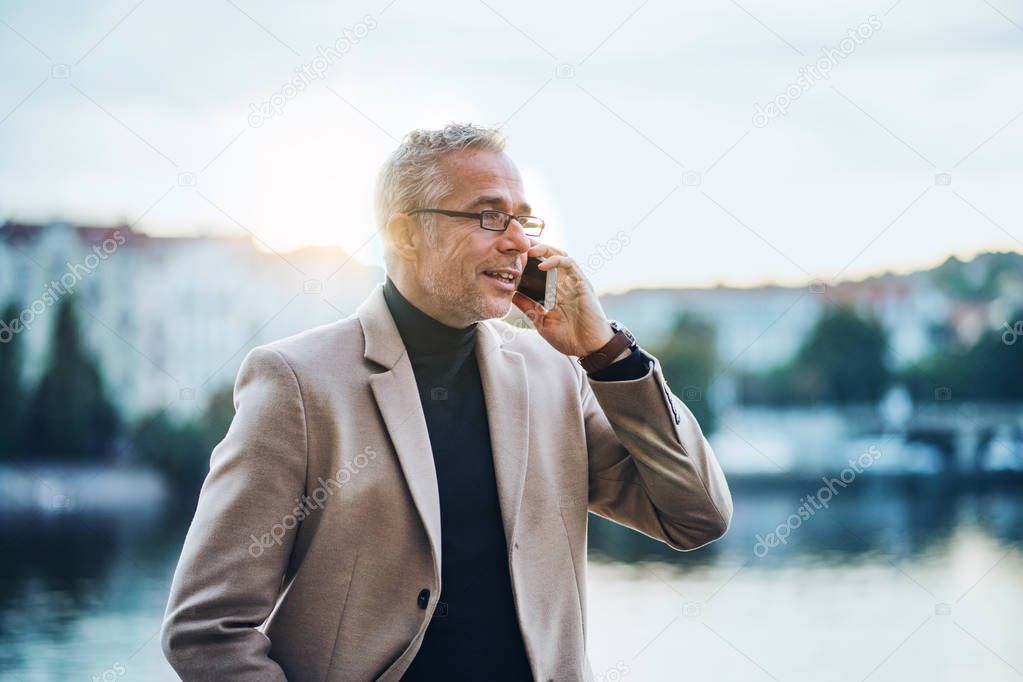Mature businessman with smartphone standing by river at sunset, making phone call.