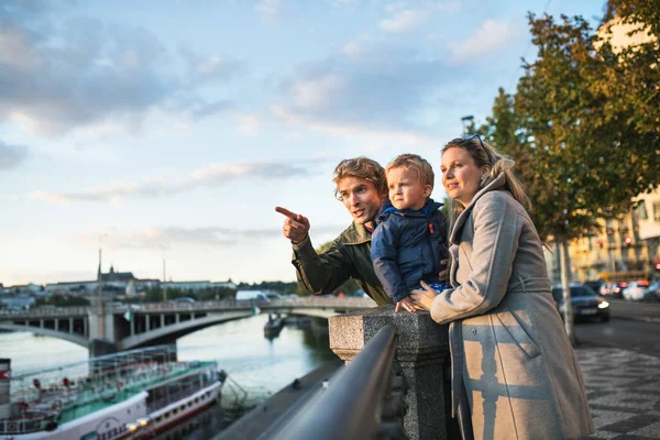 Los padres jóvenes con su hijo pequeño de pie al aire libre junto al río en la ciudad . — Foto de Stock